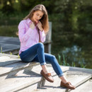 Model sitting outdoors by water wearing the pink striped shirt with blue jeans and brown loafers.
