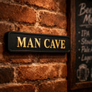 Black rectangular sign with gold lettering reading ‘MAN CAVE’ mounted on a rustic brick wall, with a framed chalkboard sign nearby.