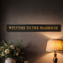 Black rectangular sign with gold lettering reading ‘WELCOME TO THE MADHOUSE’ mounted on a wall above a table with flowers and a lit lamp.