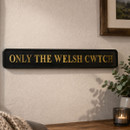 Black rectangular wall sign with gold lettering reading “ONLY THE WELSH CWTCH,” mounted on a textured cream wall above a wooden surface, with a candle, plant, and framed artwork nearby.