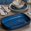 Rectangular blue ceramic baking dish on wooden dining table with plates, bowl, cutlery, and wine glass, modern kitchen table setting