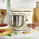 Side view of the Salter retro stand mixer with stainless steel bowl, showing a hand adjusting the speed control dial, with a finished cake and fresh berries nearby.
