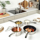 Wide kitchen scene showing stainless steel pans in use on a hob and worktop, including pasta, sauce and fried chicken dishes.