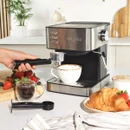 Hand holding the portafilter beside a Salter espresso machine with a cup, croissants and fresh fruit on a kitchen worktop.