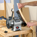 Hand guiding wood against a Draper belt sander attachment, detail sanding on a combined bench grinder in a workshop setting.