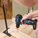Close-up of cordless drill boring into light wood plank, DXPULSE drill driver creating sawdust during DIY furniture assembly with metal hairpin legs, indoor woodworking scene.