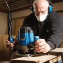 Woodworker wearing safety goggles, ear defenders and dust mask operating a Draper router on timber, focused woodworking process.