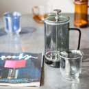 Glass cafetière on a countertop beside two clear cups and a magazine.