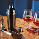 Cocktail shaker and tools on a wooden surface beside two red cocktails garnished with mint and berries.