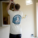 Person wearing beige Makita t-shirt installing a light fixture indoors, showing large back print graphic