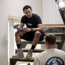 Person wearing black Makita t-shirt sitting on stairs holding a drill, indoor work setting