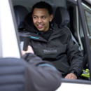 Tradesperson sitting in van wearing black Makita jacket talking and holding a coffee cup
