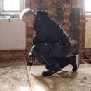 Tradesperson kneeling on a construction site wearing black Makita work trousers using a cordless drill