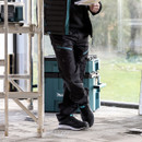 Side view of black Makita work trousers with zip pockets and reinforced knees, worn on site next to a ladder and tool boxes