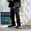Tradesperson wearing black Makita work trousers with multiple tool pockets, tape measure attached, standing on a construction site