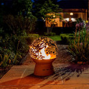 Decorative steel fire globe with intricate dragon cut-out design glowing at night, creating warm ambient light in a landscaped garden.
