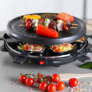 Raclette grill in use with skewers grilling on the top plate and melted cheese cooking in the individual pans below.