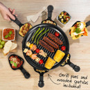 Overhead view of the raclette grill cooking skewers, sausages and vegetables, with individual pans and wooden spatulas included.