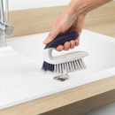 Hand using the Beldray Scrape and Scrub brush to remove dirt and grime from a kitchen sink surface.