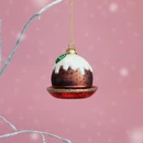 Christmas pudding bauble with white icing, green holly detail and a red base with gold glitter trim, hanging against a pink background.