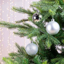 Assorted silver Christmas tree baubles with reflective, matte and glitter textures displayed on evergreen branches for festive seasonal décor.