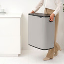 Person carrying the Brabantia Bo Touch Bin 60L in Mineral Concrete Grey using the integrated side handles for convenient movement.