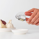 Hand using the Brabantia Tasty+ garlic press to crush a garlic clove over a small dish beside fresh garlic bulbs.