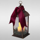 Vintage-style Christmas lantern with a bronze-effect metal frame, glowing LED candle, festive greenery and berries, finished with a large burgundy velvet bow.