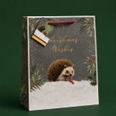 Christmas gift bag featuring a hedgehog in a snowy festive scene with Christmas Wishes lettering, foliage and red handles.