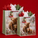 Two Christmas gift bags featuring Highland cows in a snowy winter scene with holly, red berries, festive tissue paper and red handles.