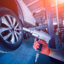 Mechanic using a pneumatic impact wrench on a car wheel in a vehicle workshop, illustrating alloy wheel nut removal with deep impact sockets.