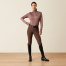 Full-length view of a model wearing a twilight mauve Ariat half-zip performance pullover styled with brown riding tights, a black belt, and tall black riding boots.