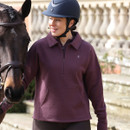 Woman wearing a thistle purple equestrian half-zip sweatshirt with a collared neckline and kangaroo pocket, standing beside a horse while wearing a riding helmet.