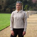 Woman wearing an oat beige equestrian half-zip sweatshirt with a collared neckline and kangaroo pocket, standing outdoors with sunglasses resting on her head.
