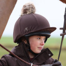 Rider wearing the Aubrion espresso riding helmet silk with faux fur pom pom while mounted outdoors.