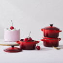 Le Creuset Cerise stoneware mini round casserole dishes styled with fresh cherries and a frosted cake, with one dish used as a serving bowl and two stacked with lids.