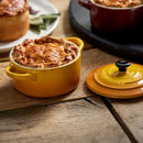 Lifestyle image of the Le Creuset Nectar mini round casserole dish filled with a golden baked pie on a rustic wooden table, with the matching lid placed alongside.