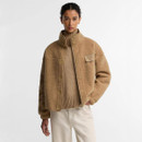 Model wearing camel teddy fleece jacket open over a tonal knitted jumper and cream trousers, showing the high collar and chest pocket.