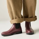 Person wearing burgundy waterproof Chelsea wellington boots with black elastic side panels and chunky grip soles, styled with rolled-up beige trousers for a casual outdoor look.