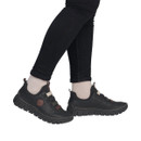 Model wearing Rieker black slip-on comfort shoes with flexible lightweight soles and decorative elastic strap detailing.