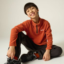 Child sitting cross-legged wearing a burnt orange half-zip fleece, black walking trousers and hiking shoes.