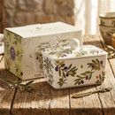 Olive Grove fine china butter dish with a fitted lid and round knob handle, decorated with a colourful olive branch pattern in green, purple and yellow. Displayed on a rustic wooden table with its matching gift box and olive sprigs, creating a Mediterranean-inspired kitchen setting.