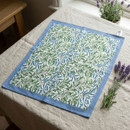 William Morris Willow Bough tea towel featuring the iconic blue and green leafy botanical design with matching blue trim and a hanging loop. Displayed on a linen-covered table alongside fresh lavender, bringing timeless heritage style to the kitchen.