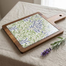 William Morris Willow Bough acacia serving board with a heat-resistant ceramic centre featuring the iconic blue and green leafy botanical design, displayed on a linen tablecloth beside fresh lavender.