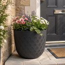 Black embossed scallop planter filled with pink, white and purple flowering plants, displayed beside a front door.