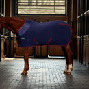 Right-side profile of a chestnut horse wearing a navy fleece cooler rug with adjustable surcingles and burgundy trim in a stable setting.