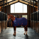 Full-body view of a chestnut horse in a navy fleece stable rug with burgundy binding, displayed in a modern stable to highlight overall fit and design.