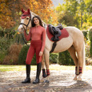 Equestrian rider in coordinated red riding wear standing beside a light-coloured horse wearing a red LeMieux saddle pad and matching fly hood.