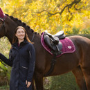 Rider standing beside a dark bay horse wearing a berry LeMieux close contact saddle pad and matching fly hood in an autumn woodland setting.