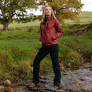 Woman in a red LeMieux hooded jacket and black trousers standing beside a stream in an autumn countryside landscape.